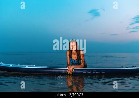 Summer sport. Portrait of young smiling tanned woman poses leaning on a sup board. Copy space. Concept of surfing. Stock Photo