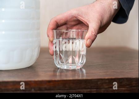 A man holds a glass of drinking water in his hand. A large white plastic bottle of fresh water is next to him on the table. Inside the room. Stock Photo