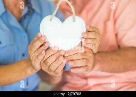 Close up of couple old hands holding white wooden heart to represent love and relationship. Heart attack prevention day life concept. Healthy unrecogn Stock Photo