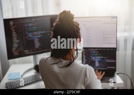 Rear of female web expert typing on computer looking at monitors while sitting indoors Stock Photo