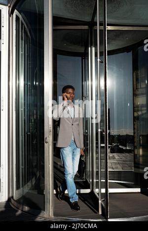 End of working day. African American manager going through glass revolving door and talking on smartphone Stock Photo