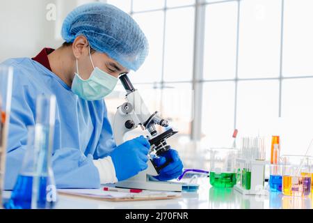 Asian male researcher in laboratory or chemist notes while studying virus sample using microscope Stock Photo