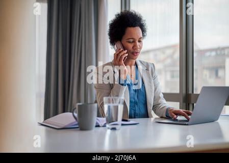 Businesswoman talking on smart phone, Female manager is working on laptop while sitting at desk, She is in formals at corporate office. Stock Photo