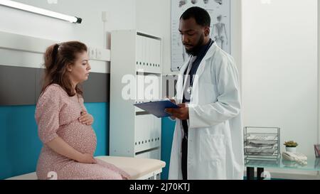 General practitioner attending consultation with pregnant woman, taking checkup notes on documents to give medical advice to future mother. Male physician having conversation with patient. Stock Photo