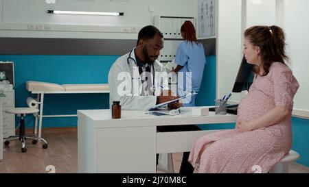 Physician consulting pregnant person and taking notes on checkup documents to give medical advice in cabinet. Male doctor having pregnancy discussion with patient expecting child. Stock Photo