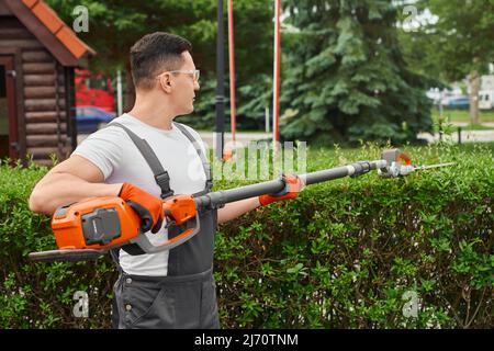 Experienced male gardener in working clothes, safety glasses and gloves using hand electric trimmer for cutting overgrown bushes. Manual work at summer garden. Stock Photo