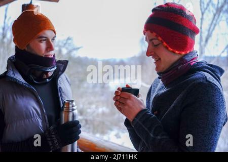 Two guys chat after skiing. They stand on the balcony of a wooden house in winter clothes, drink tea and relax. Porter of two guys in a ski resort in Stock Photo