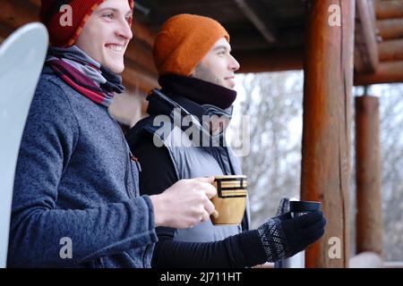 Friends chat after skiing. They stand on the balcony of a wooden house in winter clothes, drink tea and relax. Porter of two guys in a ski resort in t Stock Photo