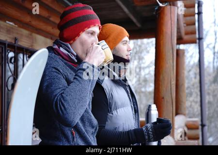 Friends chat after skiing. They stand on the balcony of a wooden house in winter clothes, drink tea and relax. Porter of two guys in a ski resort in t Stock Photo