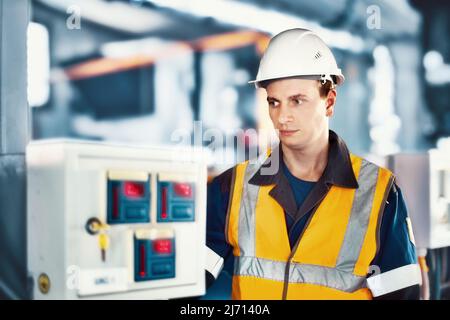 Operator or engineer in helmet and protective vest checks modern equipment in industrial plant. Portrait of Caucasian worker 30-40 years old working in production hall. Authentic scene workflow. Stock Photo