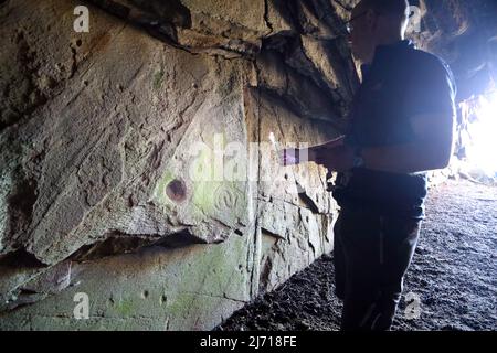 Man shining a torch illuminating the Early Christian rock carvings and possible bronze age cup markings in Scoor Cave on the Isle of Mull, Scotland Stock Photo