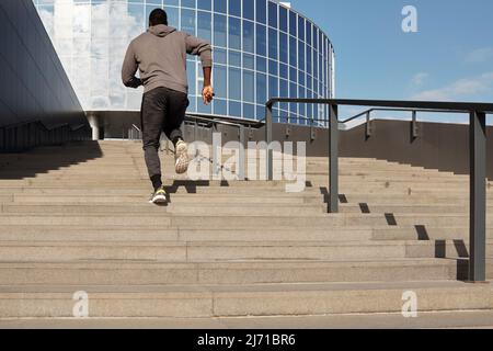 Unrecognizable Black athlete in sportswear running up the stairs towards business center with glass facade, rear view from below Stock Photo
