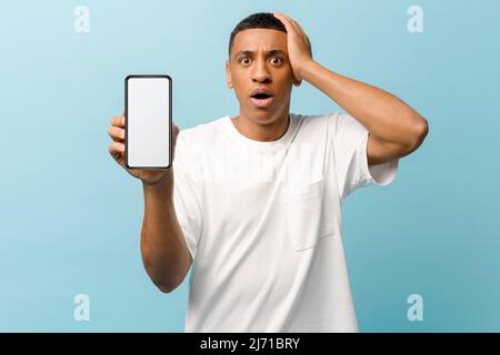Worried African-American young guy showing smartphone with blank screen standing and holding head in despair isolated on blue background, received bad notification, mockup concept Stock Photo