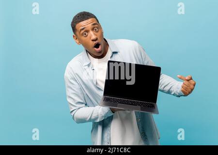 Surprised male student in casual shirt points finger at laptop pc screen with empty display isolated on blue background, shocked multiracial freelancer man dazed by possibilities of new app, mockup Stock Photo