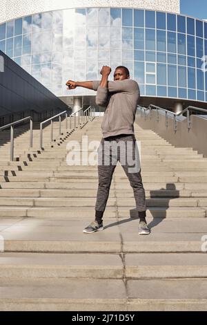 Young Black male athlete in sportswear doing warming up exercise on steps while having outdoor workout in downtown against modern office building Stock Photo