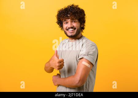 Smiling young curly Indian guy showing arm with band-aid after vaccine injection isolated on yellow background, vaccinated latino man showing thumb up Stock Photo