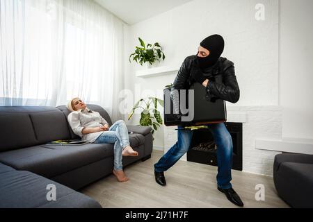 Robber man dressed in black hoodie stands with disguised face and holds a lot of money in his hands, stole a large amount, a thief man stole a TV Stock Photo