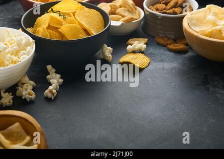 Unhealthy food. Snacks. All classic potato snacks with peanuts, popcorn and onion rings and salted pretzels in bowl plates on black concrete backgroun Stock Photo