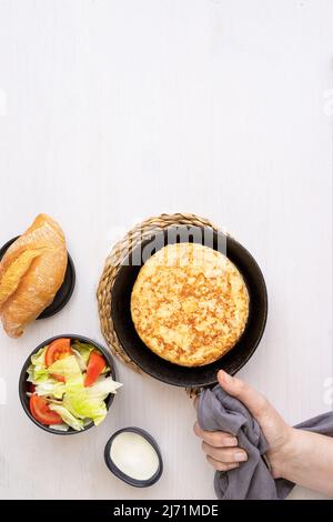 Women hand holding frying pan with Tortilla, Spanish omelette made with eggs and potatoes and served with  salad and bread. Top view. Stock Photo