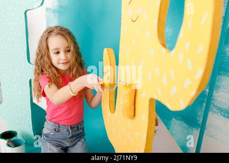 adorable little girl is doing repairs in the apartment fun paints the wall. Portrait of a beautiful smiling child with a paint roller and big paper ca Stock Photo