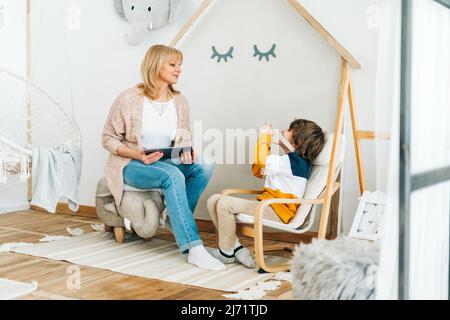 Caucasian school boy kid child taking a photo of his mother mom while she surfing the internet with a tablet. Preschooler playing with camera Stock Photo