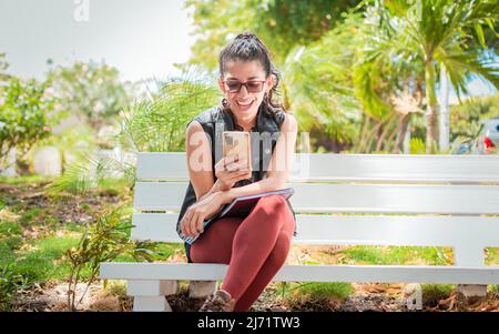 Girl sitting on a bench checking her cell phone, Happy woman sitting in a park texting on her cell phone, Woman sitting on a bench sending a text Stock Photo