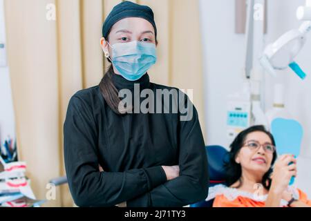 Dental doctor standing in clinic wearing mask, portrait of dentist crossing arms with patient in background, Modern Dental clinic. Dental procedures. Stock Photo