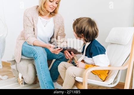 Happy caucasian grandmother sitting with grandson at home, looking in tablet computer device. Smiling family plays online video games with kid boy Stock Photo