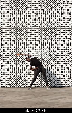 Young active Black man in sportswear doing warming up exercises before running against modern building with white perforated facade Stock Photo