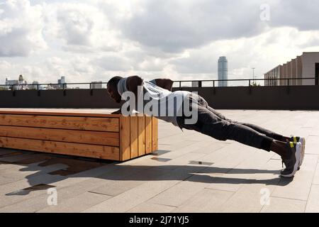 Young athletic African American man in sportswear doing push-ups against wooden bench in business park area on cloudy morning Stock Photo