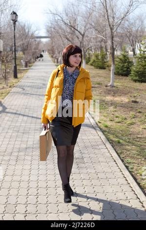 A happy 40-45-year-old woman in a yellow jacket walks down the street with a new laptop, a purchase concept. Stock Photo