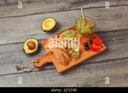 Freshly prepared guacamole in small glass bowl, bread, tomatoes, olives at working board and two avocados halves next, view from above Stock Photo