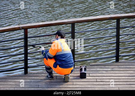 Worker in uniform paints the metal railing on a lake or river embankment. Repair works in spring park, preparing for the beach season Stock Photo