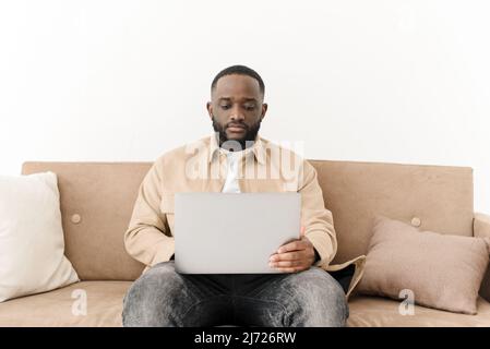 Image of focused african man working while sitting in living room. Young freelancer working from home. Stock Photo