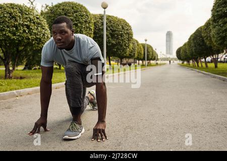 Sprinter getting ready for start. Determined athletic Black man with smartphone in armband standing on his knee on walkway in the park Stock Photo