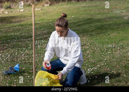 Young man crouched down to collect human pollution as plastic bags and cans in nature in a grass park Stock Photo