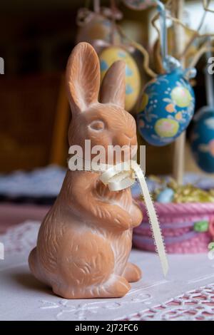 Small terracotta Easter Bunny figurine with pale yellow ribbon on neck in front of a decorative stand with Easter eggs hanging from it. Stock Photo