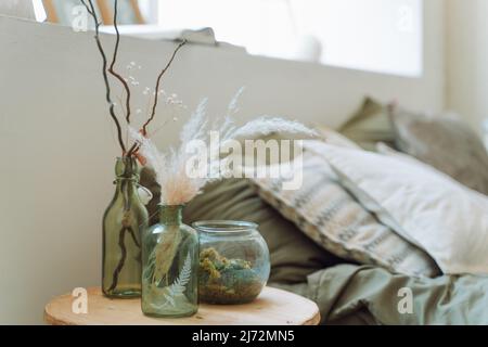 Interior of bright modern bedroom with bed covered with grey bed linen. Different green glass bottles with dry pampas, twigs, decorative plant, round Stock Photo