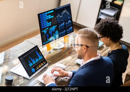 Side View Of Two Stock Market Brokers Discussing Graphs On Computer At Workplace Stock Photo