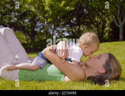 Mother laying on the grass and toddler laying on her stomach being affectionate Stock Photo
