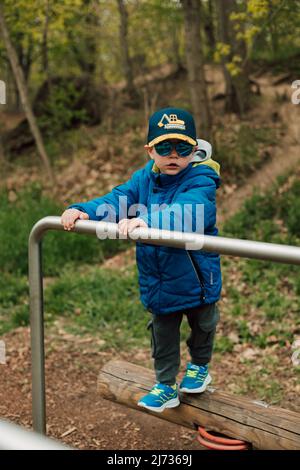 A boy in a jacket and sunglasses stands in the park leaning on the crossbar Stock Photo