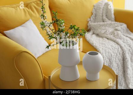 Vases with blooming tree branches on table near comfortable sofa Stock Photo