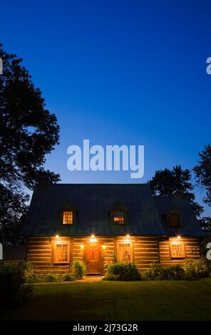 Old 1800s two-storied Canadiana style log home with landscaped front yard and grass lawn at dusk. Stock Photo