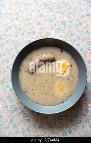 Easter sour rye soup with white sausage and egg. Traditional Polish dish for Easter. Sour rye soup in a plate and on a tablecloth with patterns. Stock Photo