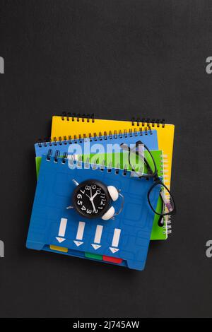 A stack of colored school notebooks and an alarm clock on a school chalkboard background. Back to school concept, banner Stock Photo