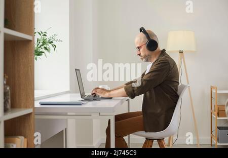 Concentrated mature man uses headphones while working on laptop computer at home. Stock Photo