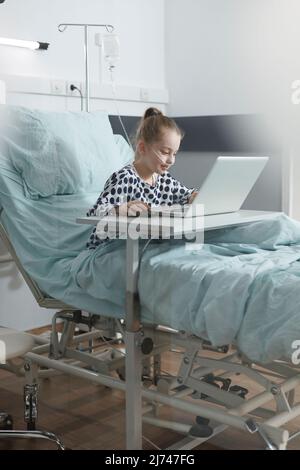 Happy ill little child enjoying video games on laptop while resting in hospital bed. Smiling sick little girl playing games on modern computer laptop while in pediatric clinic recovery ward room. Stock Photo