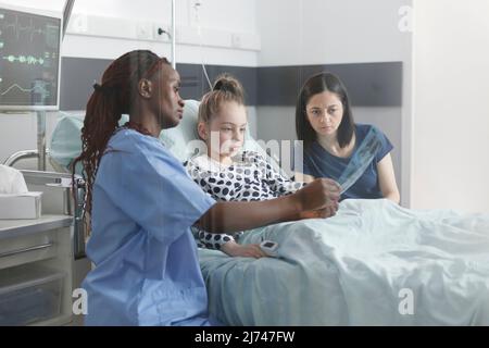 Hospital pediatric smiling nurse showing unwell little girl radiography results. Uneasy mother and surprised daughter looking at x-ray scan image while in patient recovery ward room. Stock Photo