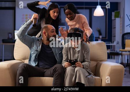 Multiethnic group of colleagues playing video games with vr glasses at office party after work hours. Sad woman losing at gameplay on console with virtual reality headset and controller. Stock Photo