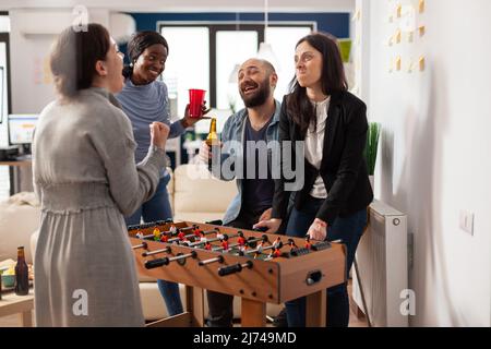 Diverse group of colleagues having fun with foosball table game, enjoying after work drinks and entertainment activity. Office coworkers celebrating after hours with alcohol and pizza. Stock Photo